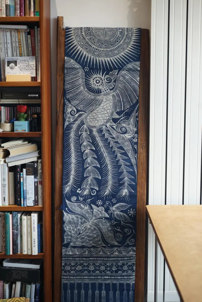 Decorative miao batik tapestry with intricate patterns hanging on a wall next to a bookshelf.