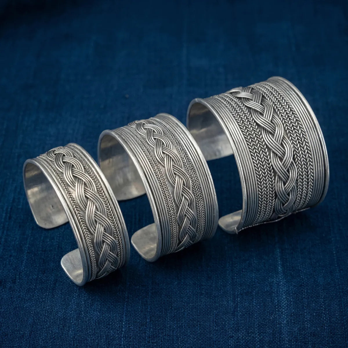 Three Miao silver bracelets with intricate patterns on a blue fabric background