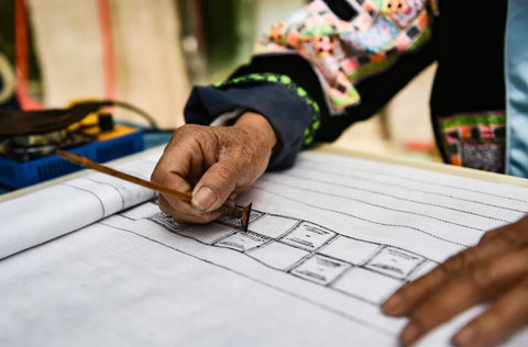 A hand is holding a wax knife and gliding across the fabric, drawing batik patterns. - Runystore