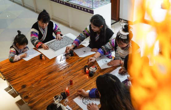 Miao artisan Cai Qun teaching young girls how to draw traditional batik wax patterns at a wooden table.