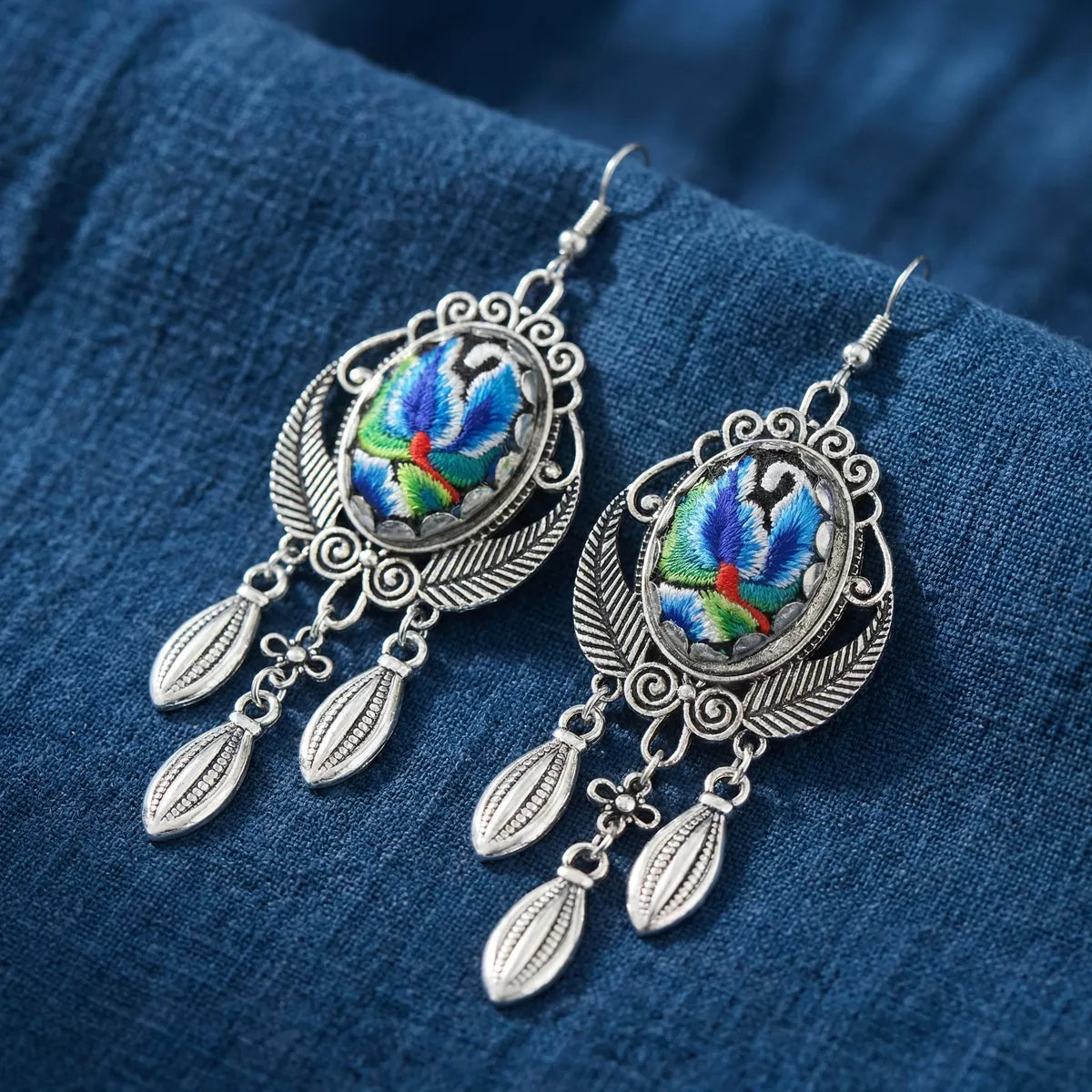 Pair of Miao silver earrings with colorful embroidered designs on a blue fabric background