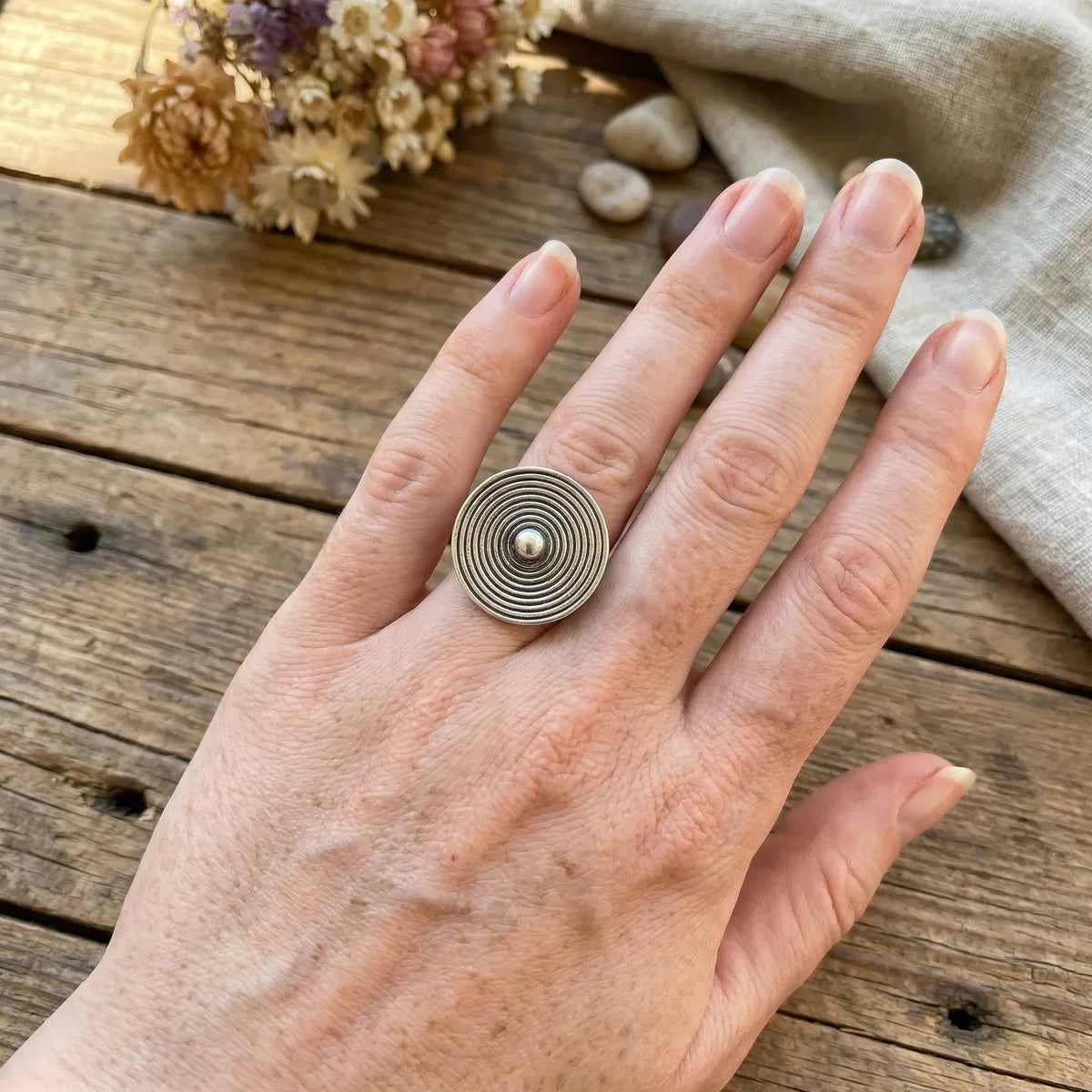 Hand wearing a miao silver ring with concentric circles on a wooden surface with dried flowers and fabric.
