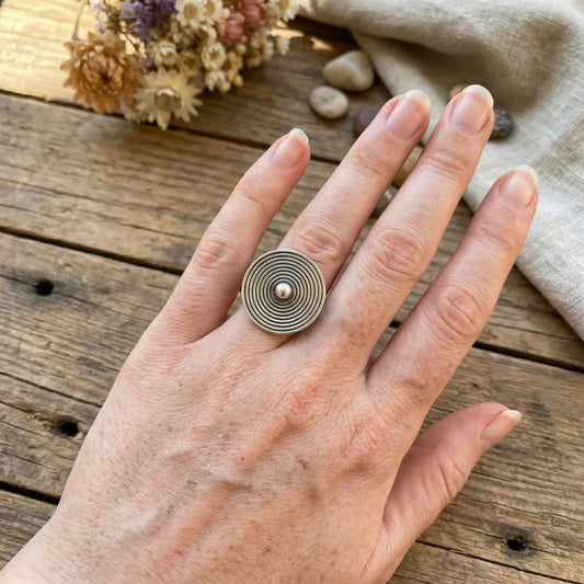 Hand wearing a miao silver ring with concentric circles on a wooden surface with dried flowers and fabric.